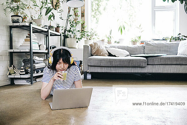 Young woman with headphones drinking smoothie while using laptop at home