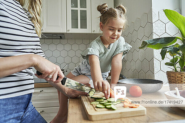 Mädchen nimmt gehackte Gurken  während Mutter in der Küche Essen zubereitet