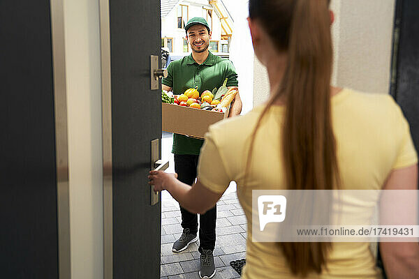 Smiling deliveryman holding vegetable box while standing at entrance