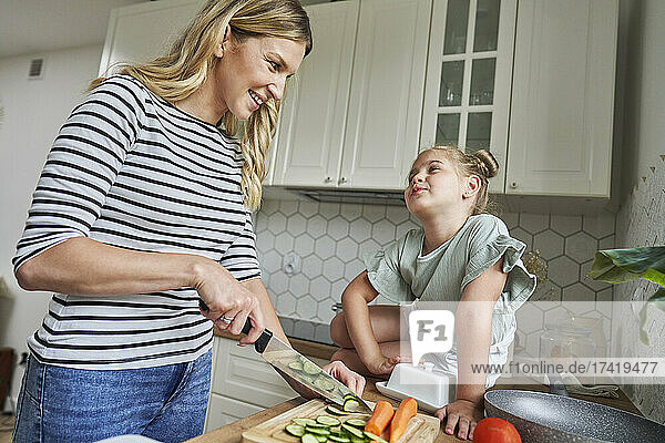 Smiling mother looking at daughter while chopping cucumber in kitchen