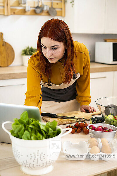 Rothaarige Frau benutzt Laptop beim Zubereiten von Speisen in der Küche