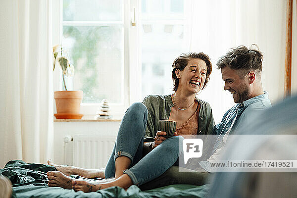 Happy woman with mug sitting with man on bed at home