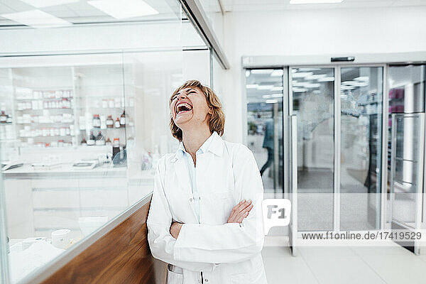 Happy female pharmacist with arms crossed leaning on glass window