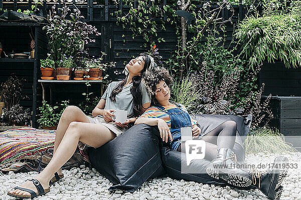 Women with coffee cups relaxing while leaning on each other at back yard