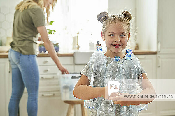 Smiling girl holding plastic bottles while mother standing in kitchen