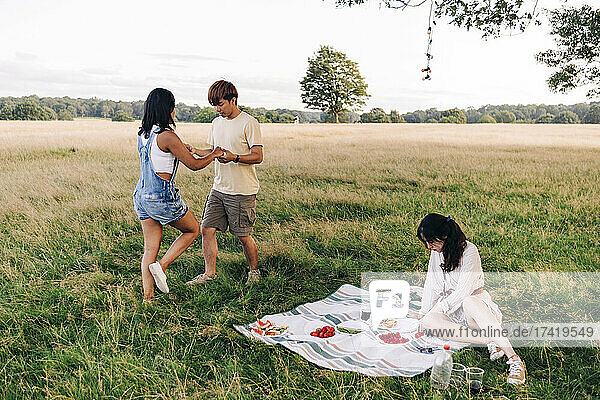 Woman sitting on picnic blanket with friends dancing at park