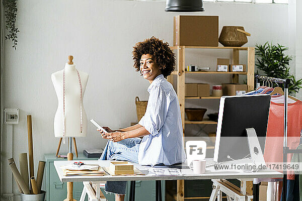 Lächelnde Designerin mit digitalem Tablet sitzt auf dem Schreibtisch im Studio