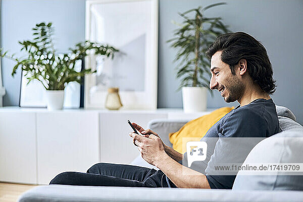 Smiling young man using smart phone while sitting on sofa at home