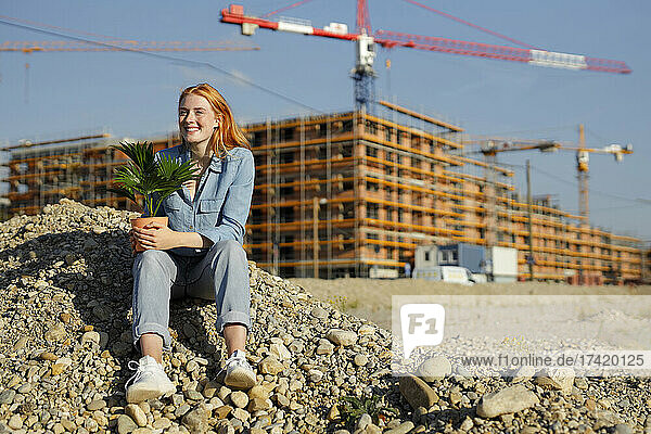 Lächelnde Frau sitzt mit Topfpflanze auf Steinhaufen auf der Baustelle