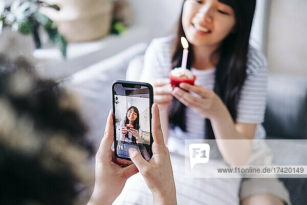 Woman photographing female friend holding cupcake through smart phone at home