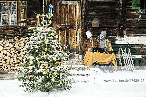 Young couple sitting with cat in front of farmhouse during winter
