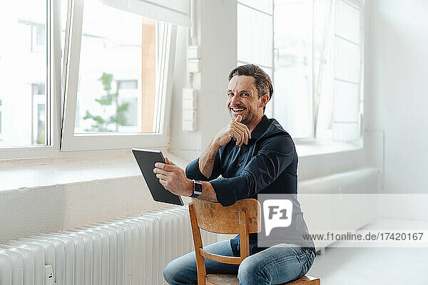 Smiling businessman holding digital tablet while sitting on chair in office
