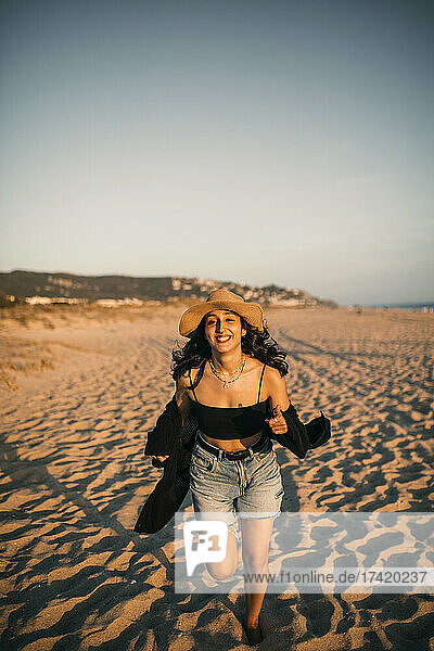 Fröhliche Frau mit Hut läuft bei Sonnenuntergang im Sand am Strand