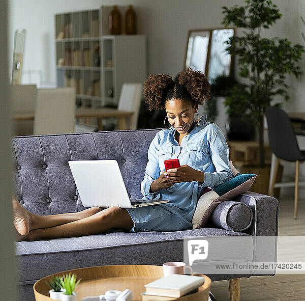 Woman using mobile phone while sitting with laptop on sofa at home