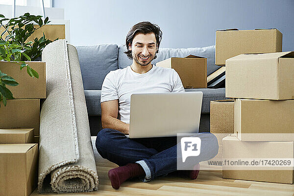 Lächelnder Mann benutzt Laptop  während er zu Hause inmitten von Kartons sitzt