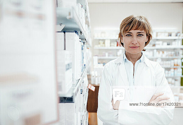 Confident female pharmacist standing by rack at medical store
