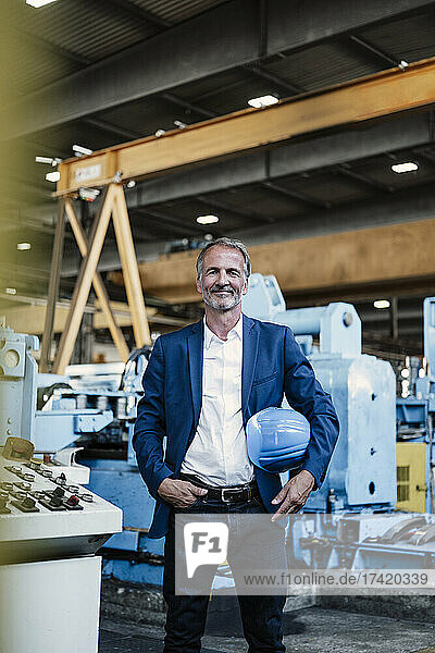 Smiling male professional holding hardhat while standing with hand in pocket at industry