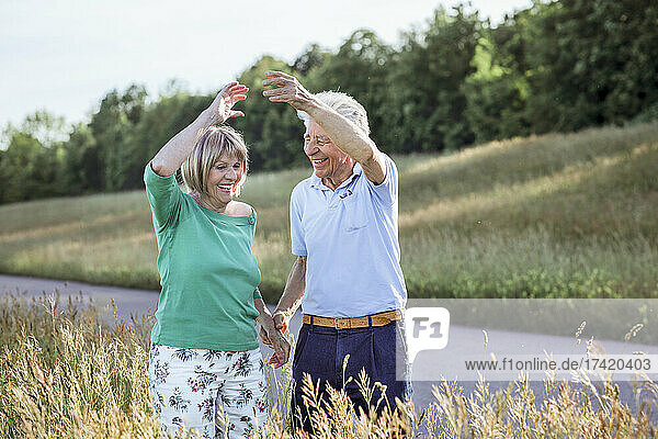 Happy senior couple with hands raised dancing while standing at meadow