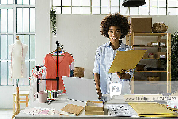 Female fashion designer with laptop holding envelope at studio