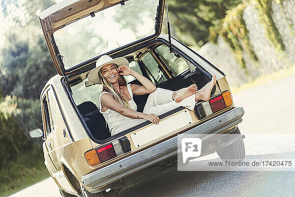 Glückliche blonde Frau mit Hut sitzt im Kofferraum eines Autos
