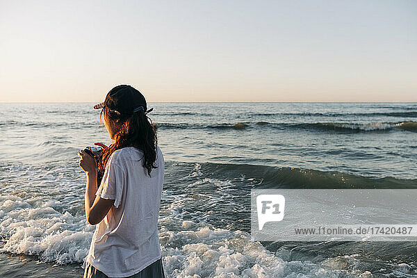 Junge Frau mit Kamera blickt im Urlaub aufs Meer