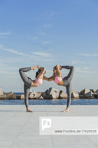 Female friends doing double dancer pose on boardwalk by sea