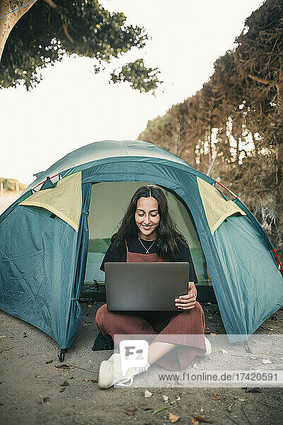 Lächelnde junge Frau  die am Wochenende im Zelt sitzt und am Laptop arbeitet