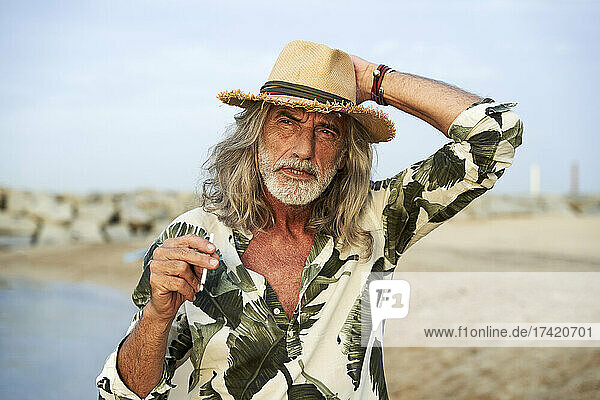 Mann passt Hut an  während er am Strand eine Zigarette in der Hand hält