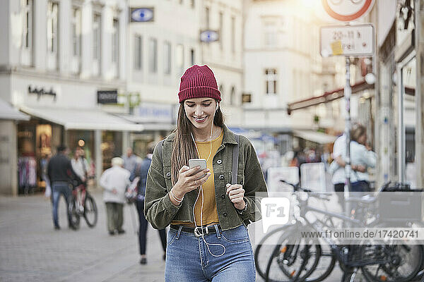 Smiling young woman using smart phone while listening music in city