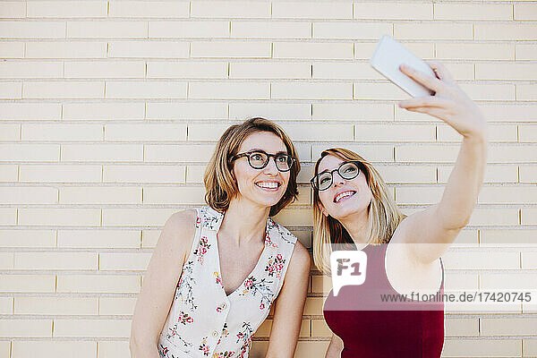 Smiling female colleagues taking selfie through smart phone in front of brick wall