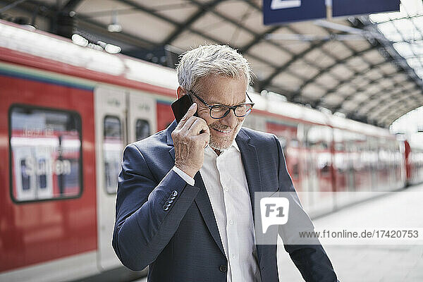 Lächelnder männlicher Berufstätiger  der am Bahnhof mit dem Mobiltelefon spricht