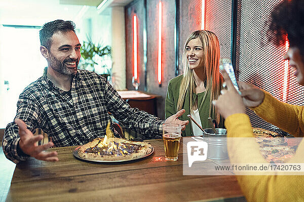 Young woman photographing man gesturing in front of flaming pizza by female friend in restaurant
