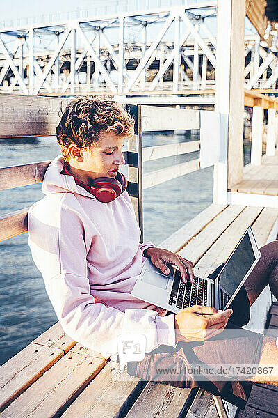 Young man using laptop while sitting on bench at pier