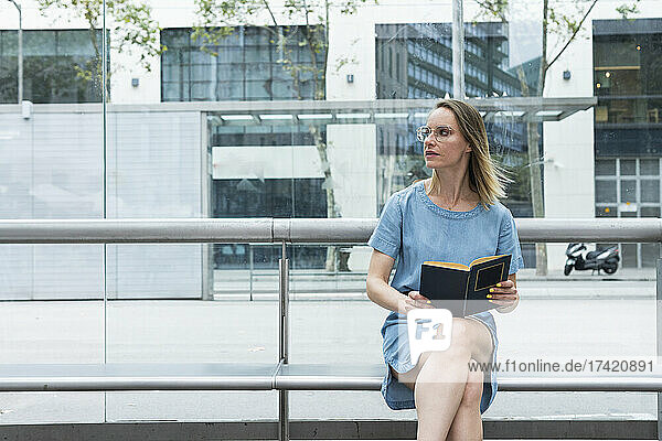 Female professional with book sitting on bench in front of glass wall