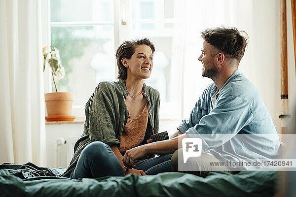 Happy mid adult couple talking while sitting on bed at home