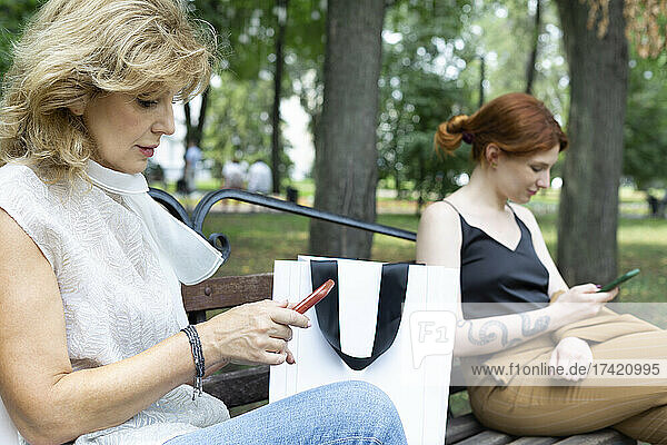Senior and young woman using smart phones while sitting on bench at park