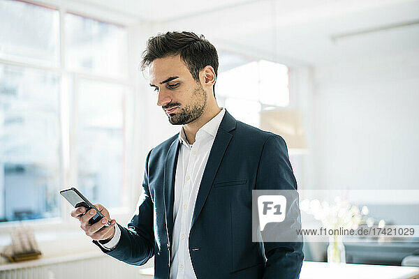 Handsome young businessman surfing net through smart phone while standing in office