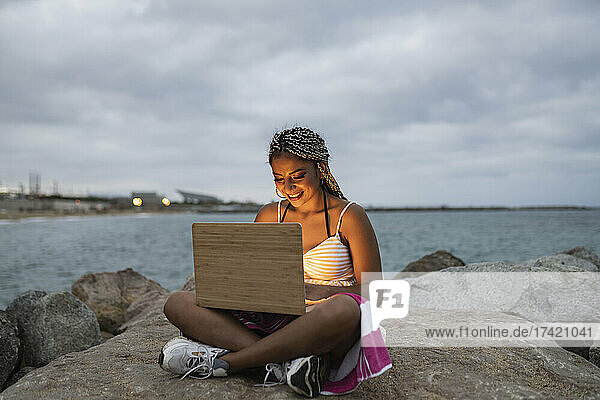 Smiling woman using laptop while sitting cross-legged on rock during sunset