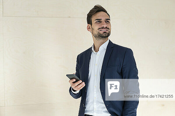 Smiling young male professional holding mobile phone while standing in front of wall