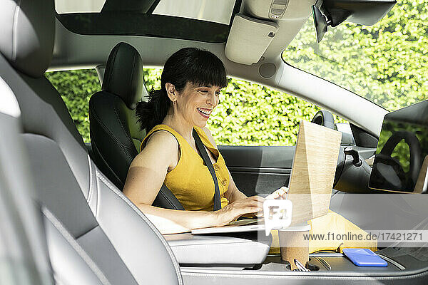 Glückliche Geschäftsfrau benutzt Laptop  während sie im Elektroauto sitzt