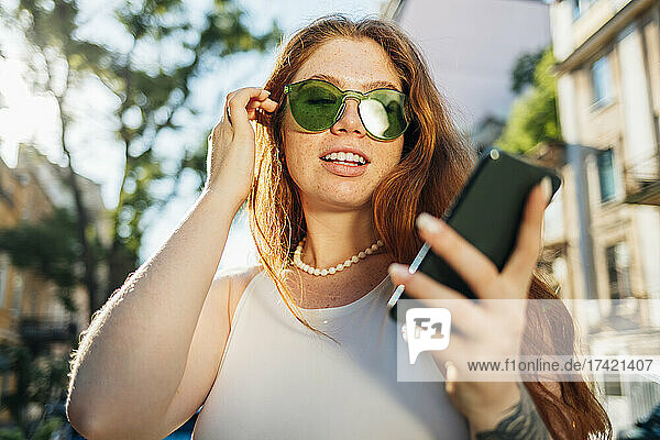 Young woman with hand in hair wearing green sunglasses using smart phone