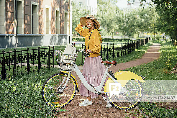 Smiling woman standing with bicycle on footpath in public park