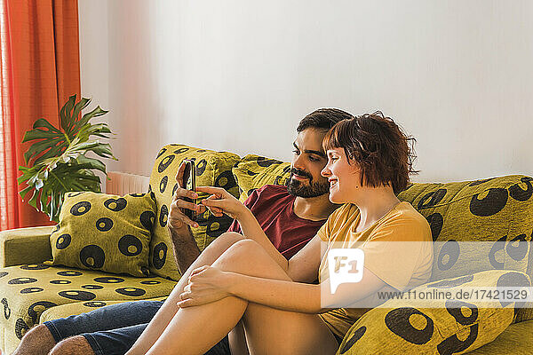 Bearded man sharing mobile phone with girlfriend while sitting on sofa in living room