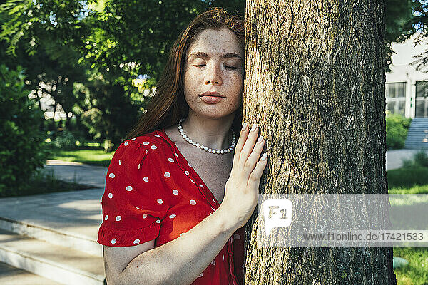 Young woman with eyes closed standing by tree trunk in park