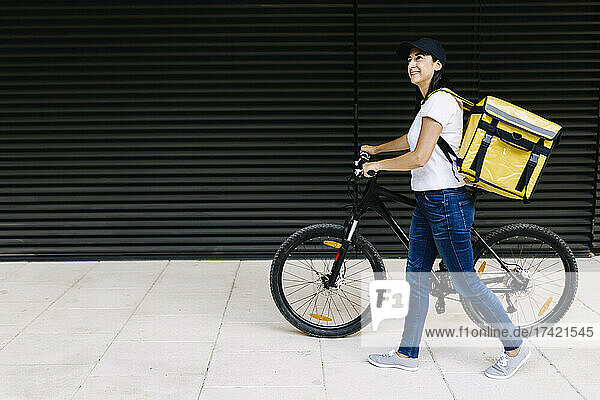 Smiling delivery woman walking with bicycle on footpath