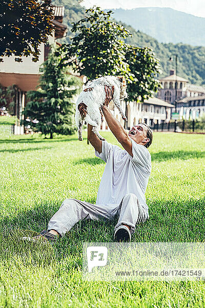 Glücklicher Mann hebt Hund hoch  während er im Gras sitzt