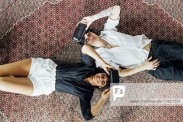 Smiling lesbian couple removing virtual reality simulators while lying on carpet at home