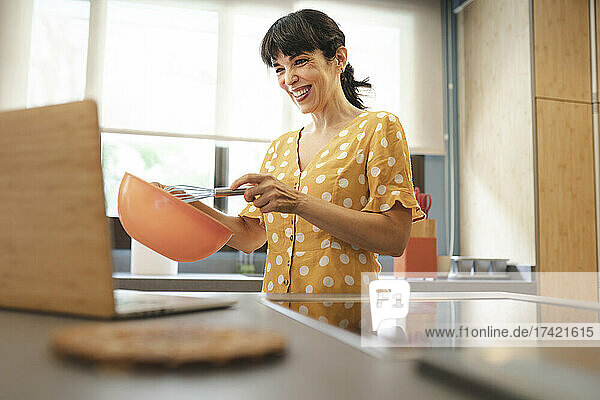 Smiling mature woman preparing food while learning through laptop in kitchen at home