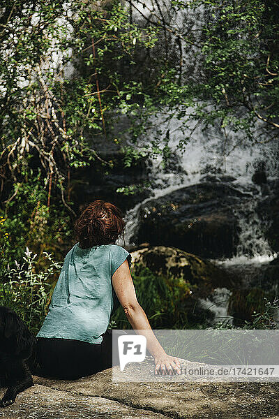 Mid adult woman looking at waterfall while sitting with dog in forest