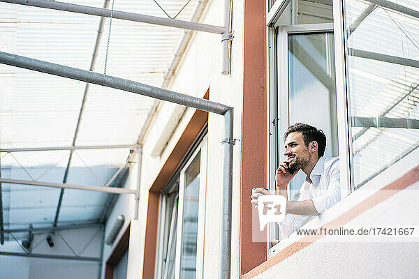 Smiling young businessman holding coffee cup while talking on smart phone at window